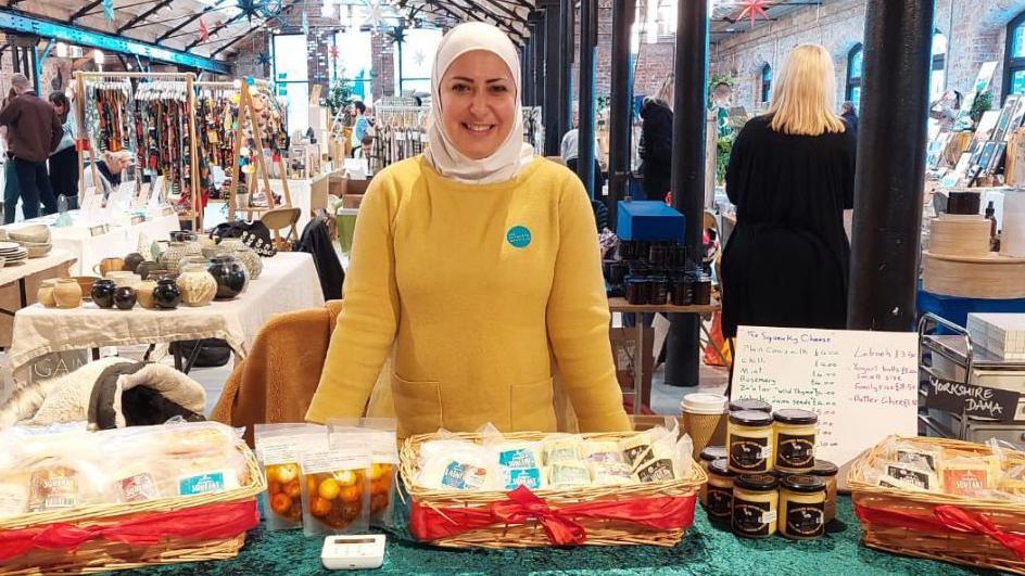 Razan Alsous wearing a yellow top and white head scarf standing behind a stall of various cheeses in an indoor market. Other stalls are in the background, with shoppers looking at items for sale. 