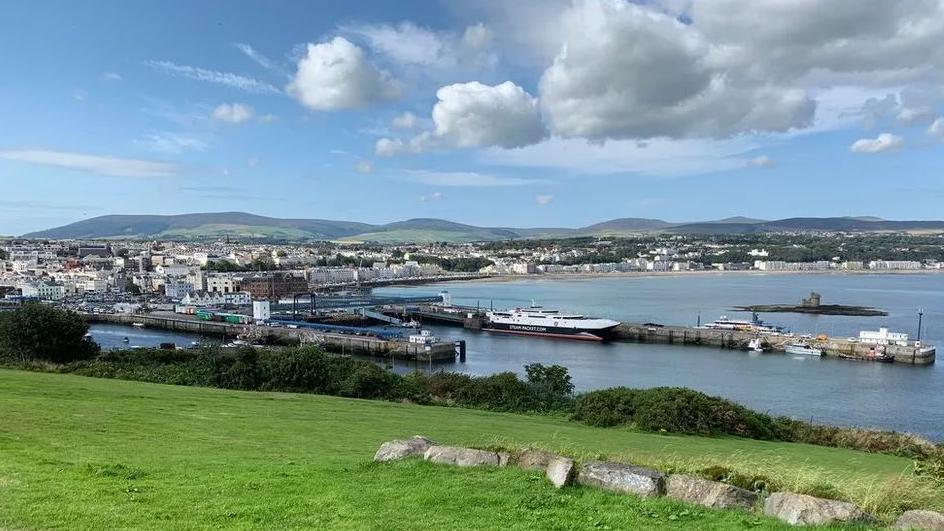 A view of Douglas Harbour, with green grass in the foreground and blue skies. You can see the Tower of Refuge on an islet in the bay and the fastcraft Manannan alongside a pier. 