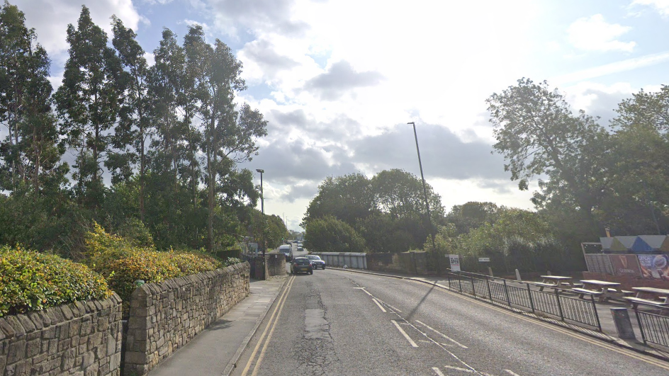 A street with a 5ft brick wall on one side and railings on the other with a bridge ahead, and tall trees to one side.