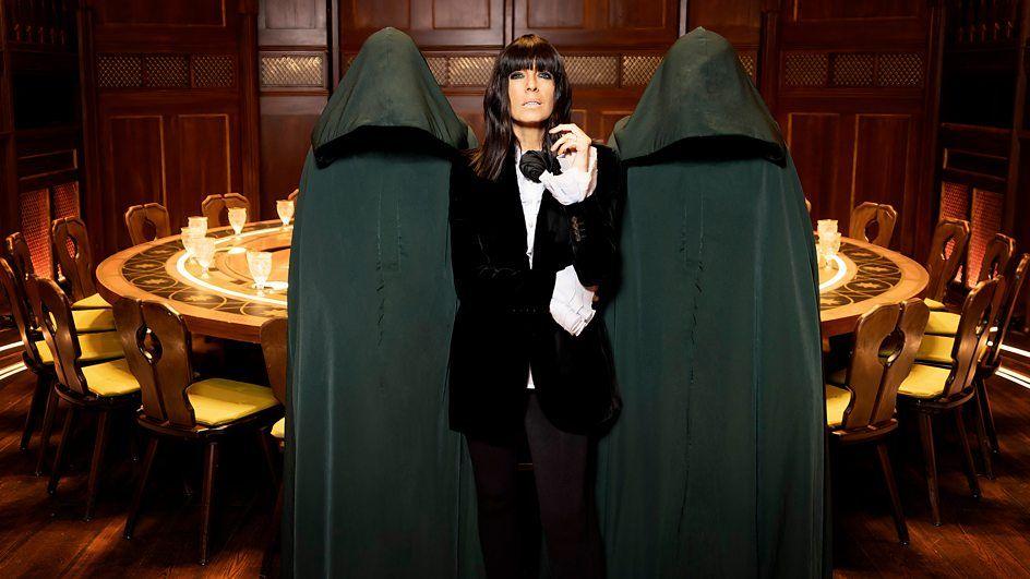 Claudia Winkleman, in a black velvet suit, with white shirt and black bow-tie in the shape of a rose, posing in between two people whose faces and bodies are hidden under full-length black cloaks. In the background is a dark wood panelled room containing a large round-table and chairs.