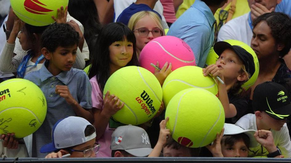 Children holding giant tennis balls wait for the end of the women's singles round of 16 tennis match between USA's Coco Gauff and USA's Emma Navarro to receive autograph