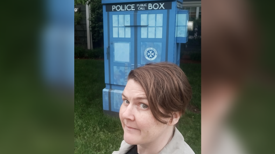 Erica with short dark hair, wearing a black t shirt and beige blazer, stood in front of the Tardis