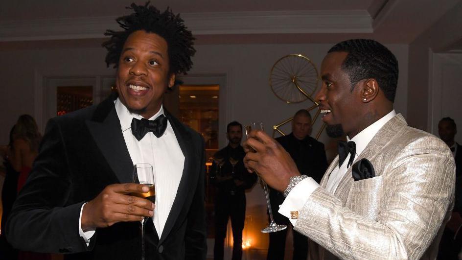 File image of Jay-Z and Diddy at an event - Jay Z is dressed in black tie and is smiling and looking off to his right, while Sean Combs, on the right, is looking across at him and smiling. He is wearing black tie, but with an shiny ivory coloured jacket. Both men are holding champagne flutes. 