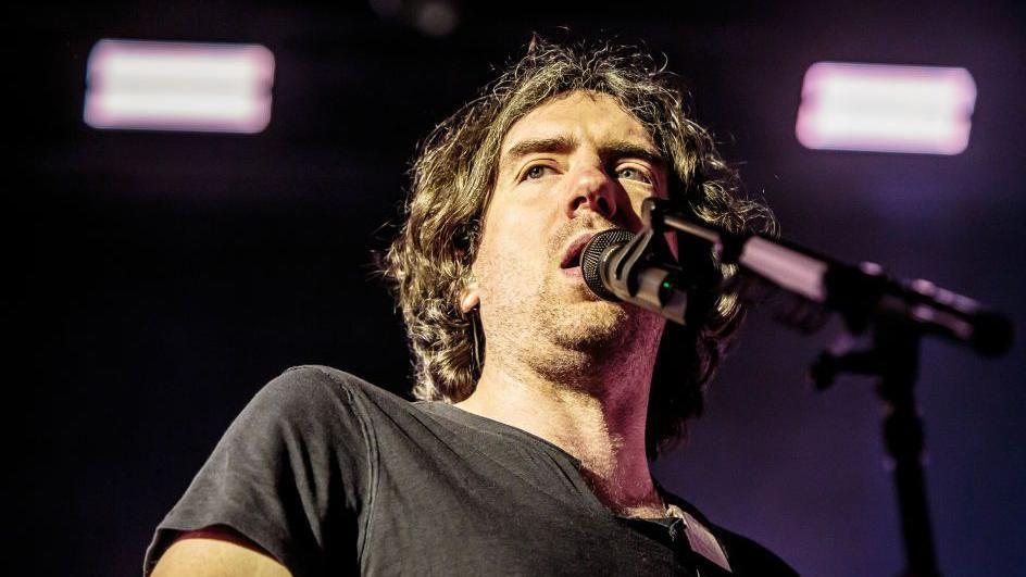 Gary Lightbody in dark t-shirt, with long dark wavy hair, about to sing into microphone.