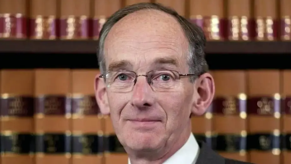 A man with short grey hair and glasses looks at the camera with a neutral expression. He is facing the camera with shelves of books behind him.