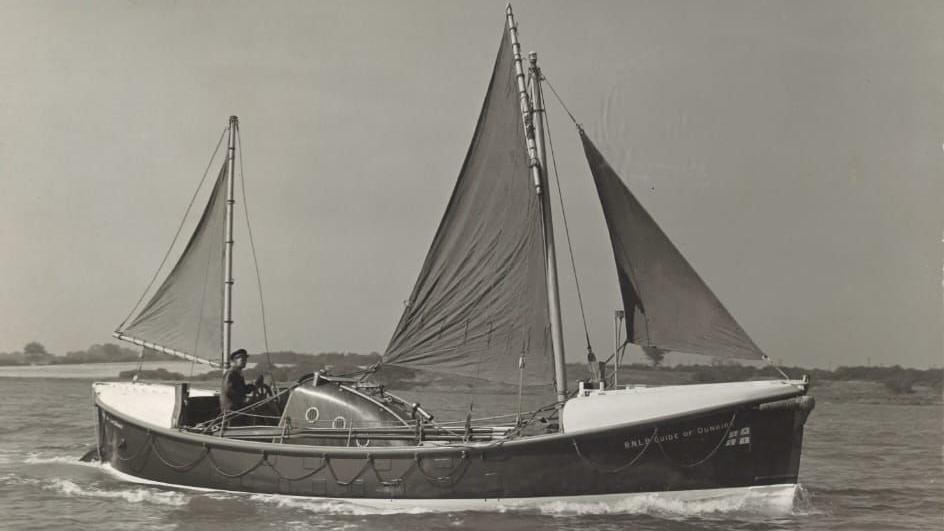 A black and white image of the Guide of Dunkirk navigating through water. A man is steering the vessel. The vessel has three sails.