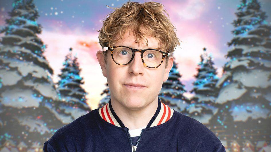 Josh Widdicombe wears glasses and a navy blue sport jacket with a red and white trim collar. 