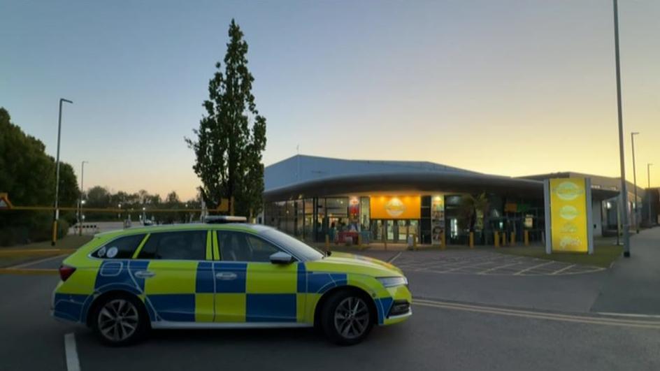 A blue and yellow police car stationary outside a building with an orange and blue sign that reads "Waterworld". It is dusk and the sun is setting in the sky