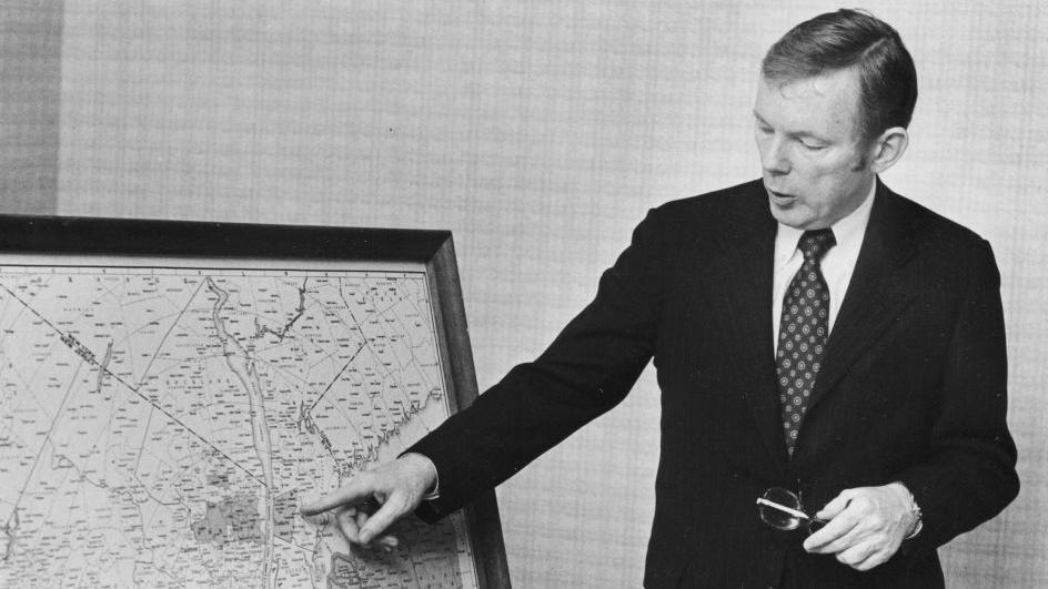 Black and white photo of Charles Dolan in a dark suit pointing to a map of New York