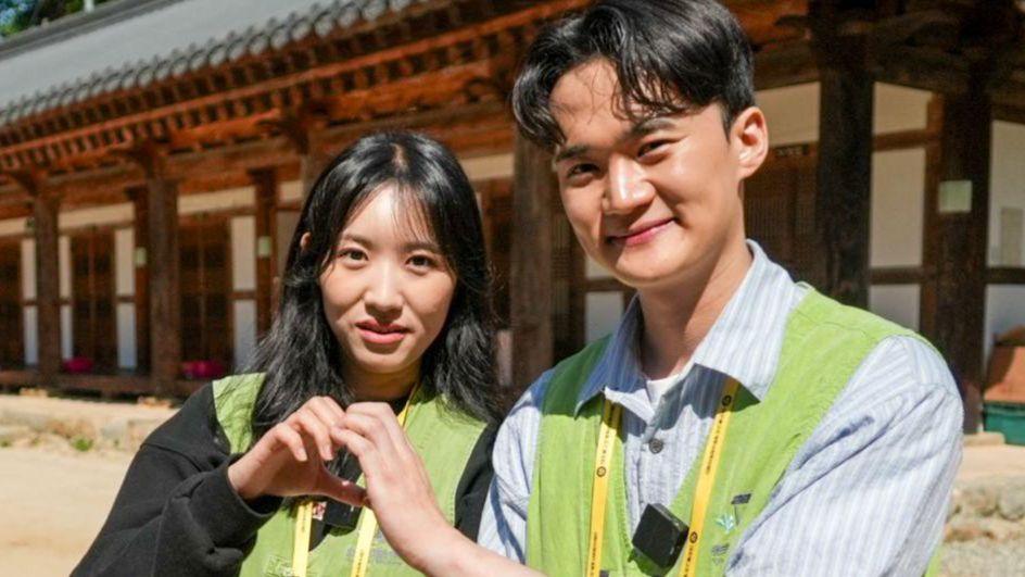Two participants - Ruby and Minho - wearing light green vests and event lanyards stand in a courtyard, joining hands to form a heart shape.