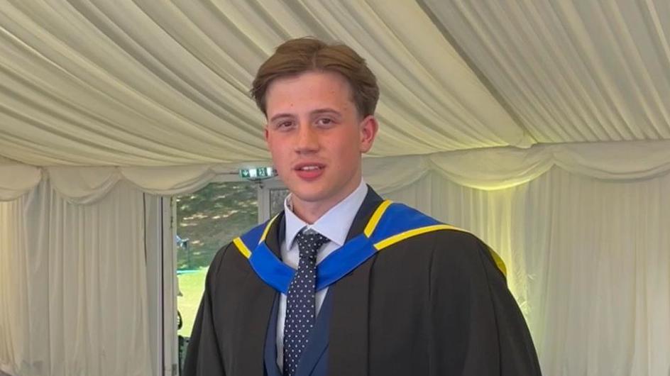 James Harrison at his graduation, he's standing inside a cream tent. He's wearing a black graduation gown with blue and yellow accents. 