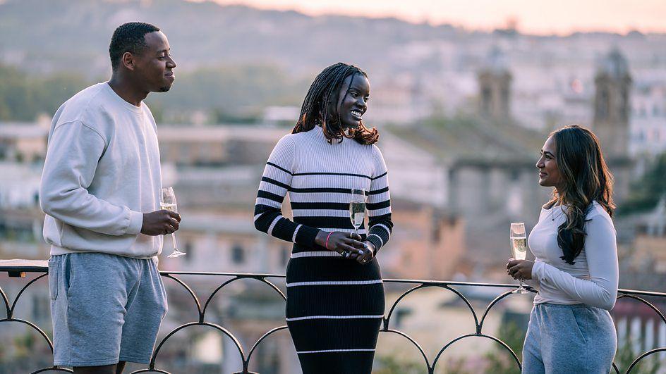 Three people are standing next to each other by a railing overlooking a city. They are looking at each other and smiling while holding champagne flutes.