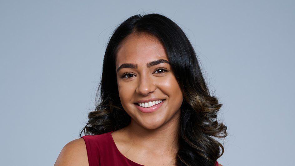 A young woman with curled hair smiles at the camera. She wears a maroon top and stands in front of a grey background.