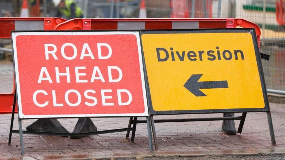A red road closed sign is placed on a city street, beside a yellow sign advising people of a diversion. Both signs sit on a red brick surface