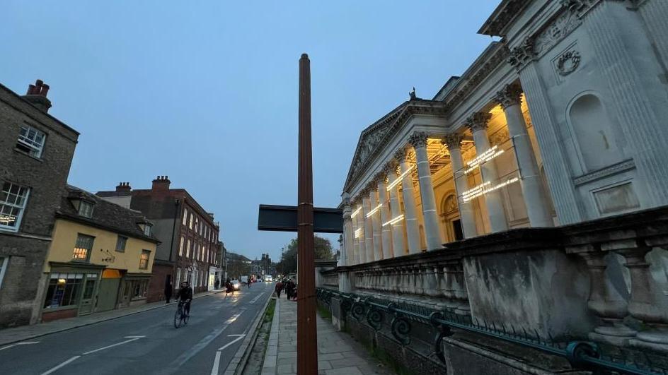 Street view showing the front of the Fitzwilliam Museum lit up behind its ornate columns. On the path outside is a post which is missing the lamp on top. There is a cyclist riding along the damp road outside the museum