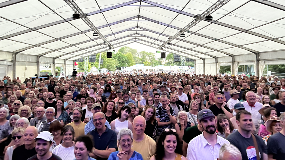 A large crowd of people all facing the stage at the folk festival. The camera has been positioned on the stage so all of their faces can been seen. They have gathered under a large white tent and many people are smiling. 