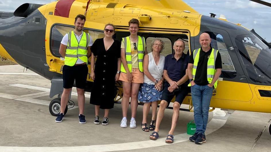 Six people in front of an air ambulance. Three are wearing high-vis jackets and are all looking at the camera. Joe and his wife, Muriel, are sitting down, the other four are standing up. The ambulance is black and yellow in colour.