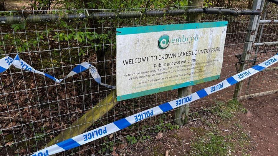 A metal fence with a Crown Lakes sign board on it and blue and white police tape around it.