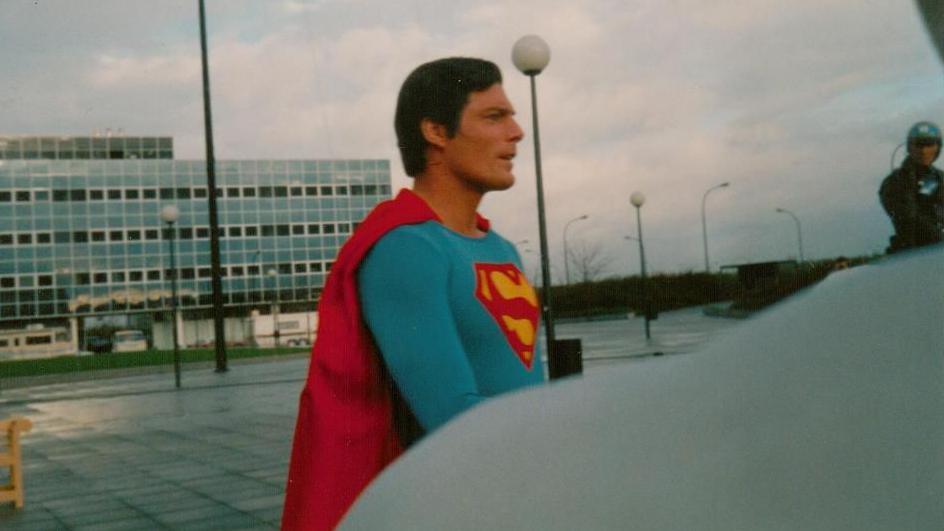 Christopher Reeve in a Superman costume stands outdoors in front of a modern building with many windows. The sky is cloudy and there are street lamps are in the background.