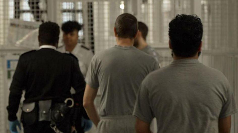 Men wearing prison clothing are escorted in a line by a uniformed prison officer inside a prison in north London