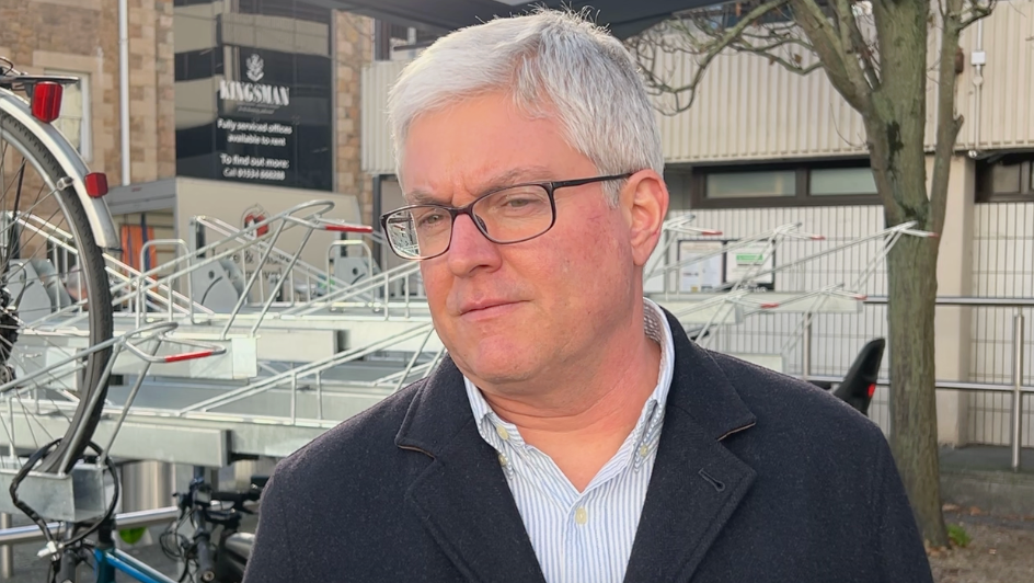 A man standing outdoors near a covered bike shelter that contains several bicycles and a multi-tier bike rack. In the background, there is a building with signage that reads “KINGSMAN”. The setting appears to be an urban area with paved ground, metal railings, and a tree without leaves. The man is wearing a dark coat over a light collared shirt.