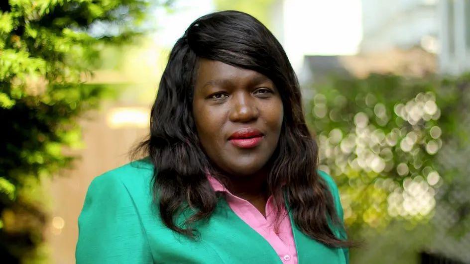 Head and shoulders image of Florence Nyasamo. She is in a teal green jacket and pink shirt. She is long brown/black hair and the background is green, but blurred.