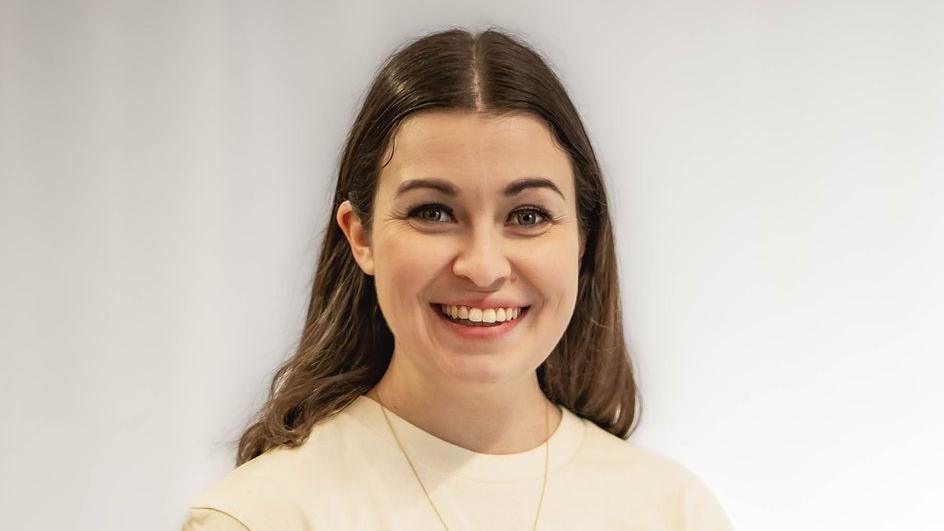 Lauren Troiano standing by a wall, wearing a white top. She has long dark hair and a necklace on, and is smiling.