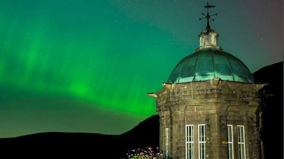 An old looking green domed building in the foreground with a green Northern lights sky in the background 