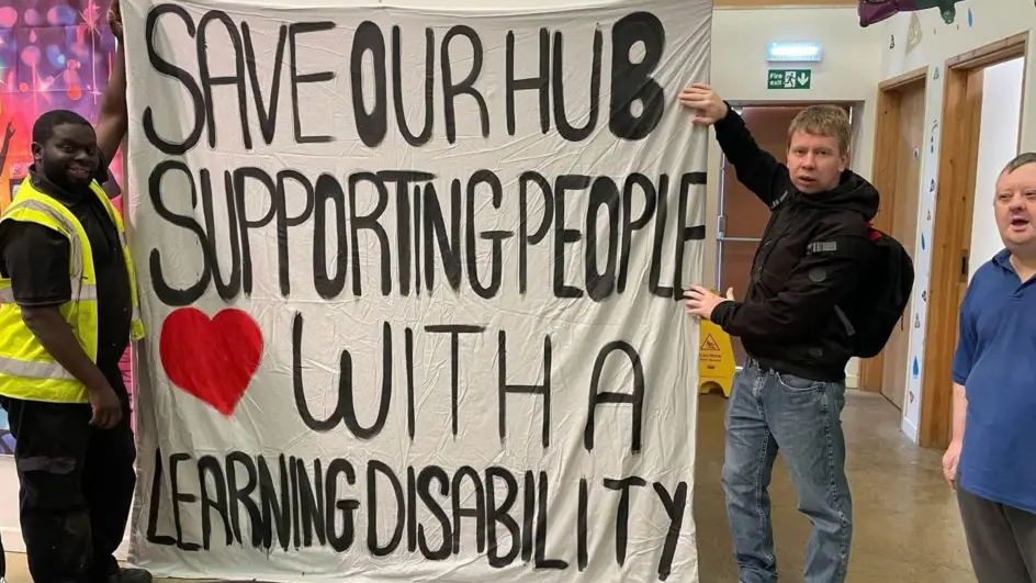 Two men are holding a large banner indoors, and a third man is in the far right of the photo. One man is dressed in black wearing a high-vis yellow vest, and the second man posing with the banner is wearing a black coat and blue jeans. The banner reads: "Save our hub supporting people with a learning disability".