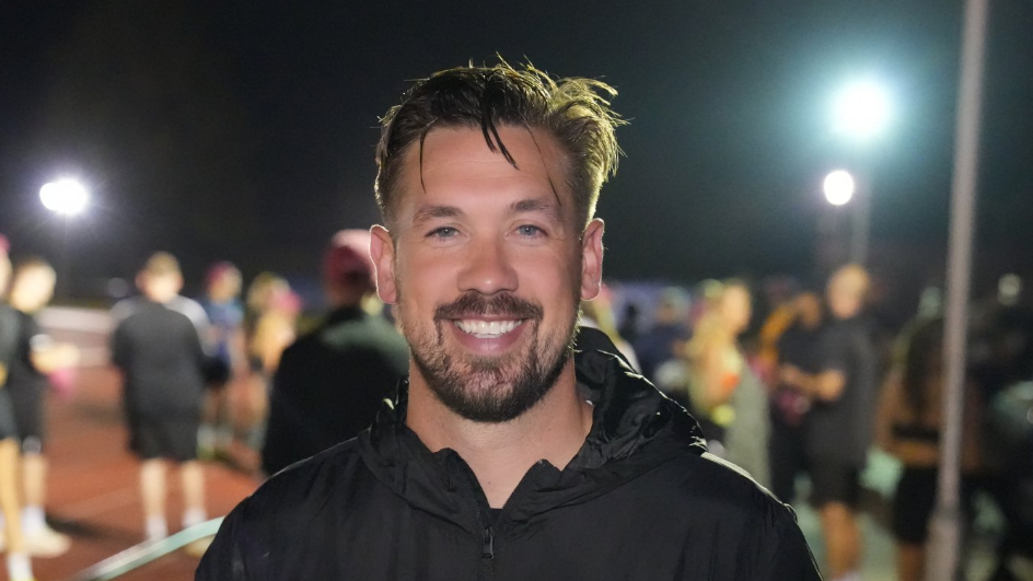 Luke Chambers is smiling into the camera. He is wearing a black sports jacket. Behind him are other runners on an athletics track. 