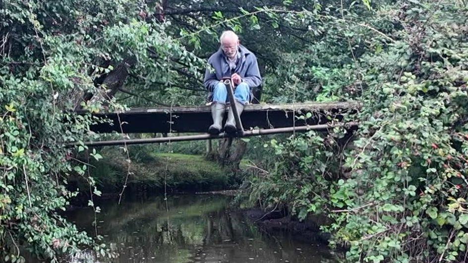 Mike James sitting chained to a small footbridge crossing a stream with foliage on both sides