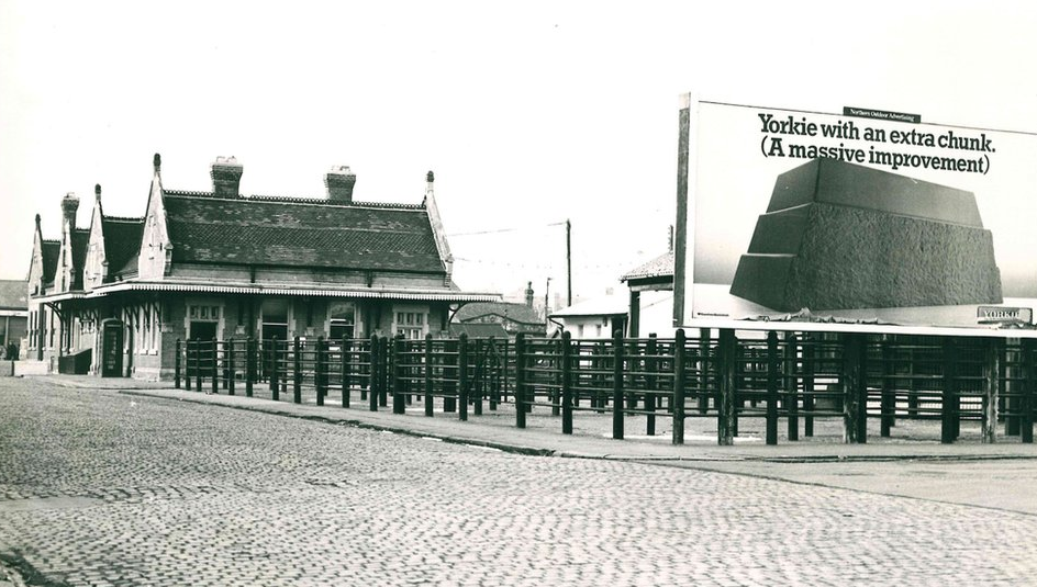 A black and white photo showing livestock pens in a cobbled area in front of counting house buildings. A large advertising board for Yorkie chocolate is on the right of the image.