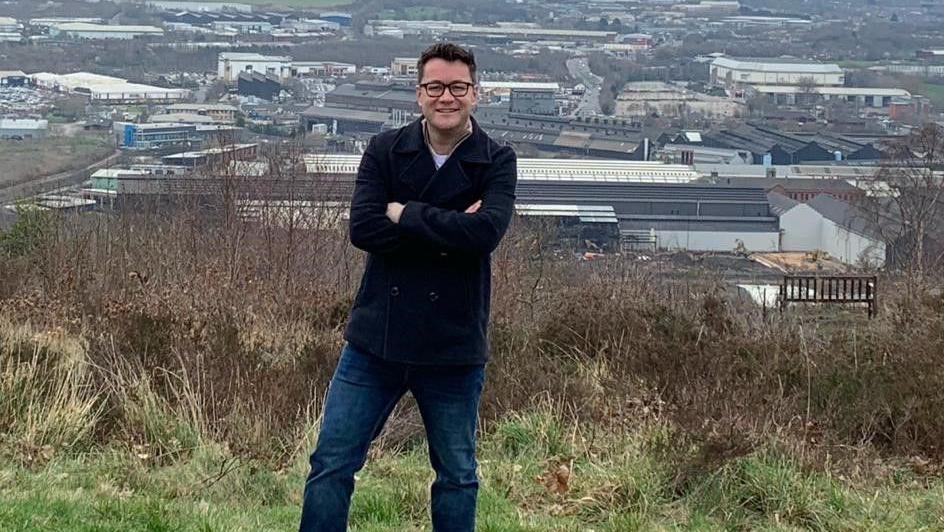 Mark Rusling, a dark-haired man wearing glasses, and dressed in jeans and a navy coat, is pictured standing on a hill with Sheffield in the background.