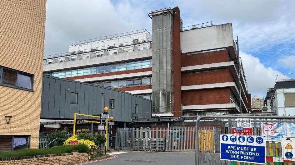 Four-storey concrete hospital building in the background with a first-floor walk way to a modern brick building in the foreground. Metal fencing with a PPE warning is also visible.