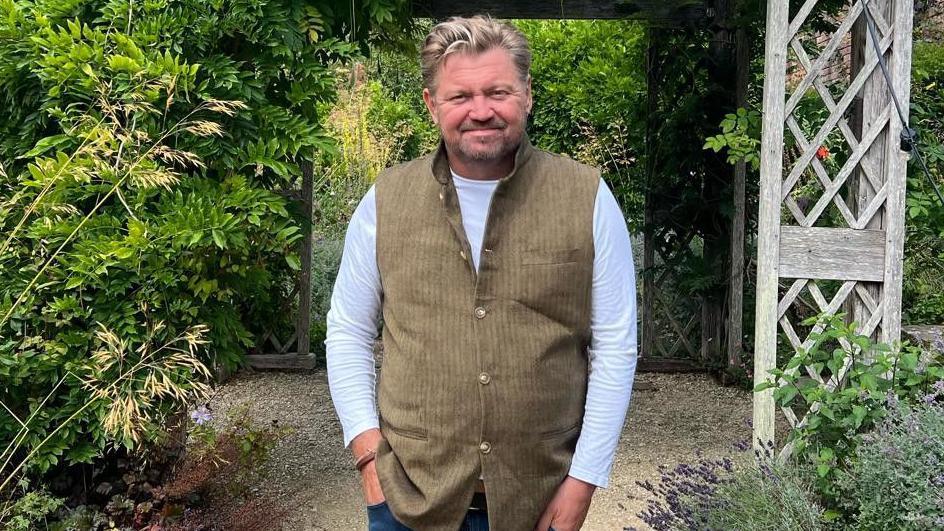 James Perkins stands smiling in a garden. He has a beard and combed-back fair and grey hair and wears a brown jerkin over a long-sleeved white T-shirt.