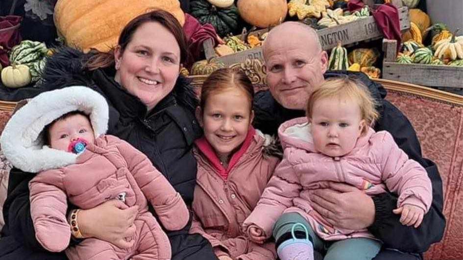 Mike and Laura Barlow, both wearing black coats, hold three young girls, all dressed in pink, at a Halloween event.