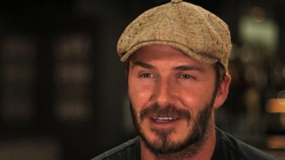 David Beckham wearing a sandy coloured flat cap and dark T-shirt.