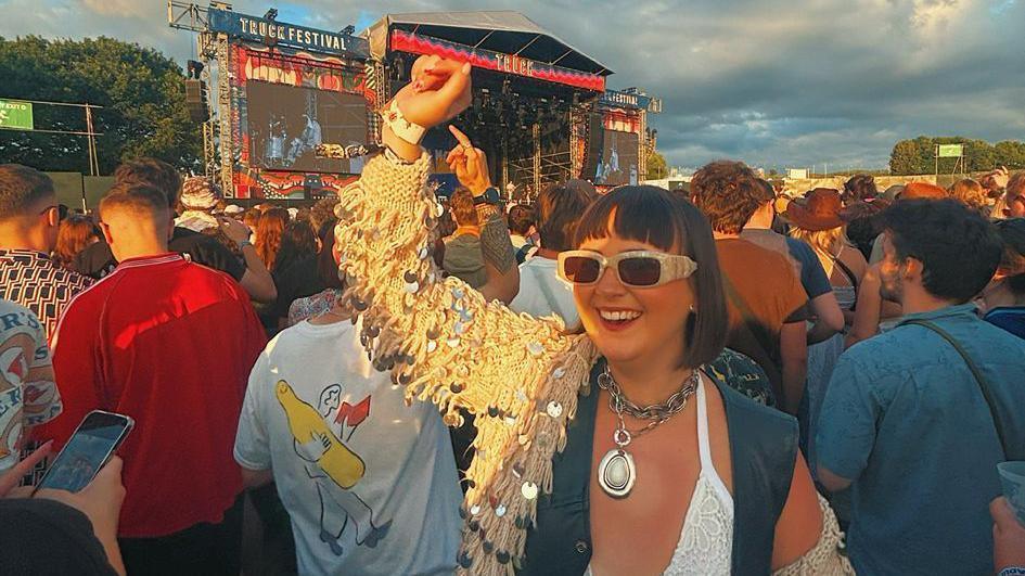 Lucy stands in a crowd in front of a stage which says Truck Festival on it, surrounded by people looking away from camera at the stage as she looks towards it, smiling with her arm in the air. She has short brown hair with a fringe over her forehead, and is wearing a sparkly tasseled jacket.