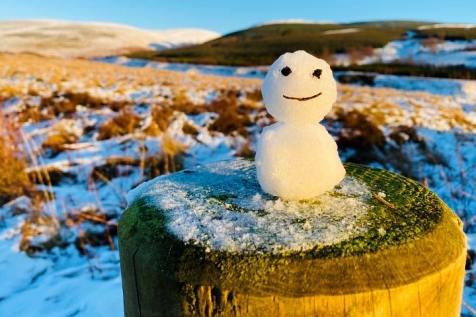 The snowman is sitting on a fence post. It has two eyes and small twig for a smiley mouth. Moorland behind the scene is covered in snow.