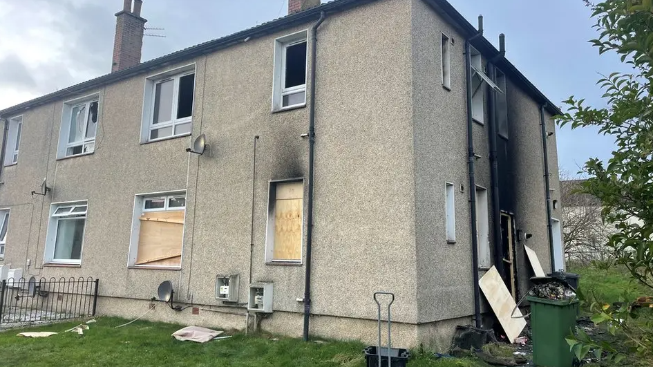 A general view of a block of flats in Auchinleck. The stone is light. Several of the windows are boarded up with light wooden chipboard. The side of the building has a large black scorch mark the length of the building.
