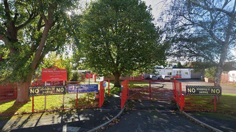 The outside of a primary school. The school has red gates and fences, and three large trees on the grass.