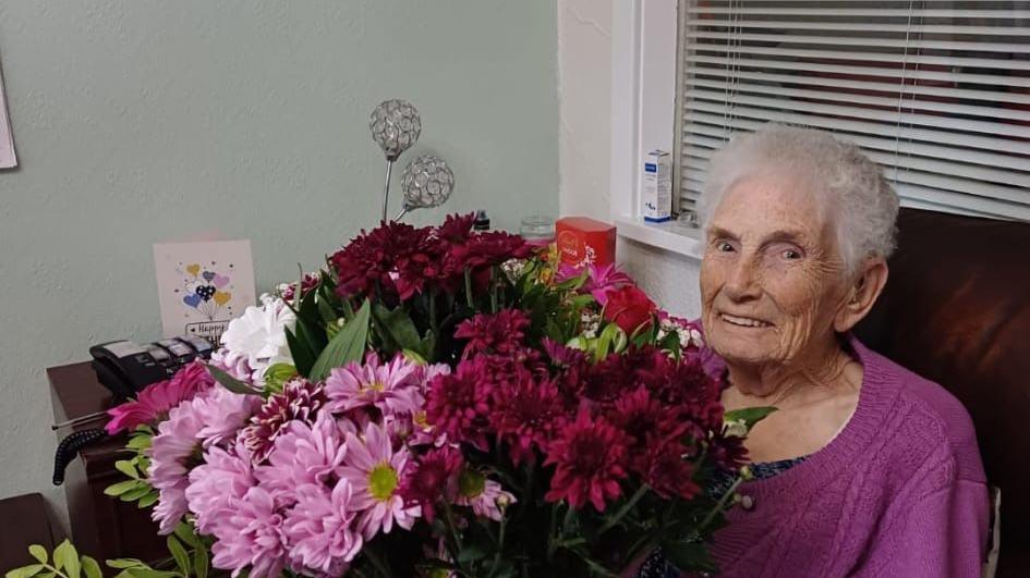 Lilian smiling at the camera, she is in a purple jumper and is holding birthday flowers. There are pink, red and white flowers. 