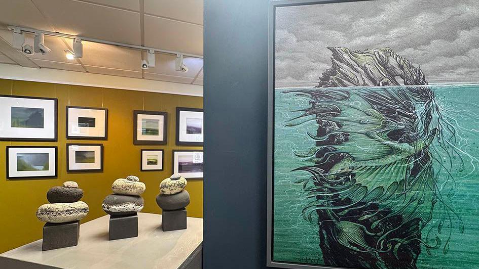 An exhibition, featuring an illustration of a green dragon under water with its wings wrapped around a rocky piece of land that has a rocky top that is protruding above the water line. In the background there are other landscape paintings and clay sculptures.
