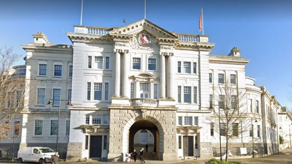 The outside of Kent County Council county hall. The building is three storey's high and made of brick. Lots of the brick is painted white. At the centre of the building is an archway. 