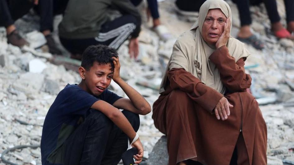 A woman wearing a brown robe and beige head cover with a hand to the side of her face looks distraught as she sits next to a boy wearing a navy blue T-shrt and dark trousers with the left hand on the side of his face and head, crying at a funeral.