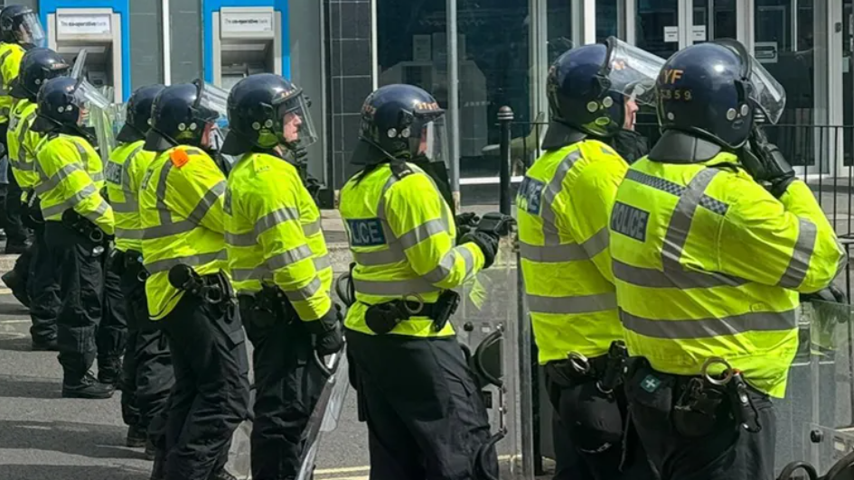 A line of police officers in riot gear, wearing helmets and holding shields