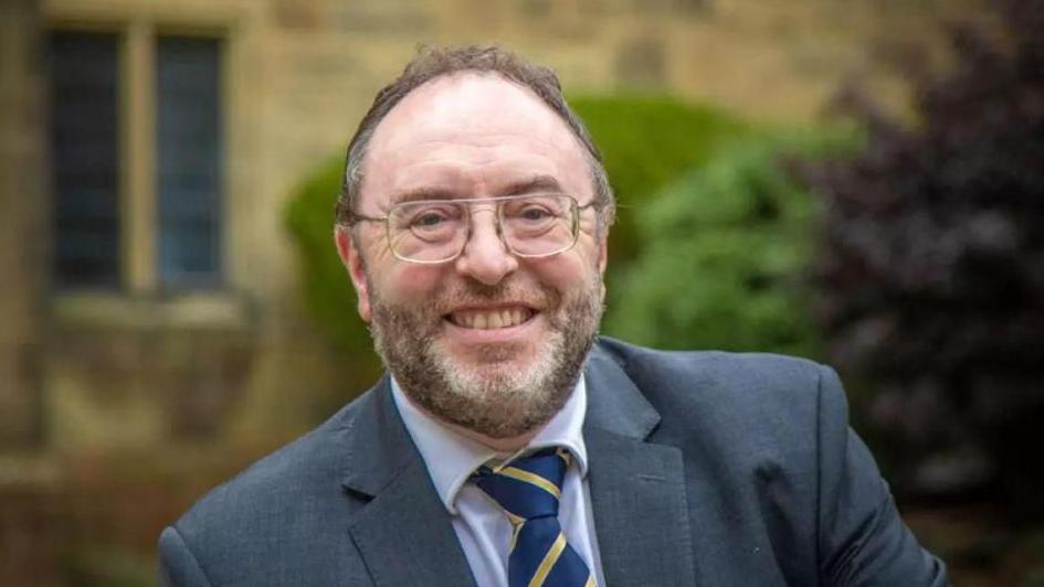 Edmund Burke smiles at the camera. He has short brown hair and a beard slightly greying. He is wearing square-framed glasses and a grey suit jacket. A white shirt and a blue and yellow stripe tie. Behind him a blurred background of a historic yellow brick building.