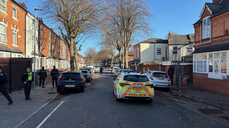 A police car can be seen parked in the middle of the road in front of police tape set up in two areas cordoning off the scene. Cars are parked on either side of the road, outside houses and officers can be seen in the street.