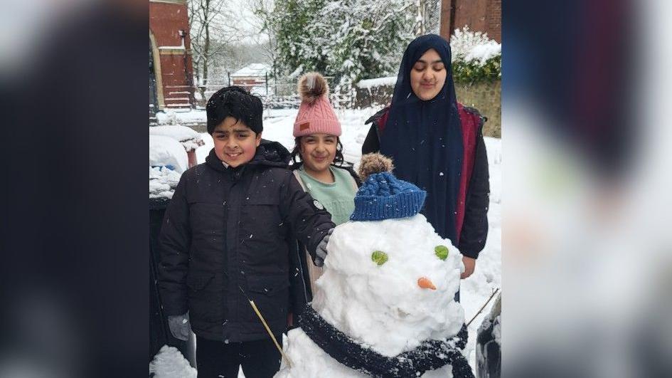 A boy and two girls stand next to their snowman, who has a carrot nose and sprouts for eyes, while wearing a blue bobble hat.