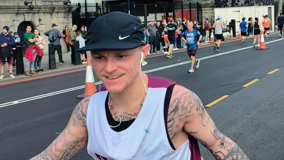 Jack Taylor running the London Landmarks Half Marathon last year. He is wearing a black and white top and has a baseball cap and headphones. There are crowds on the side of the road.